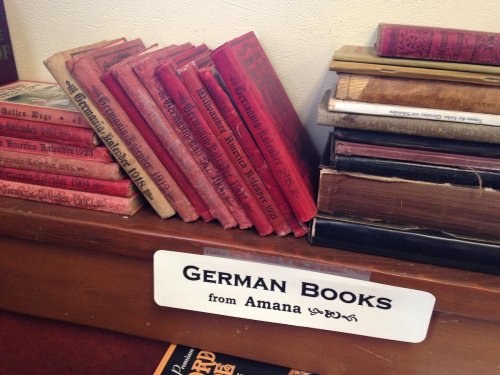 A collection of vintage German books stacked on a shelf, with a sign reading "German Books from Amana."