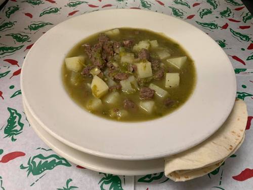 A bowl of green chili stew with beef and potatoes, served with a side of tortilla on a patterned tablecloth.