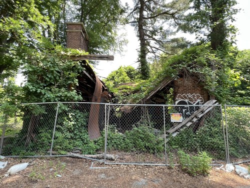 Abandoned building covered in vines and graffiti, partially collapsed, surrounded by a chain-link fence.