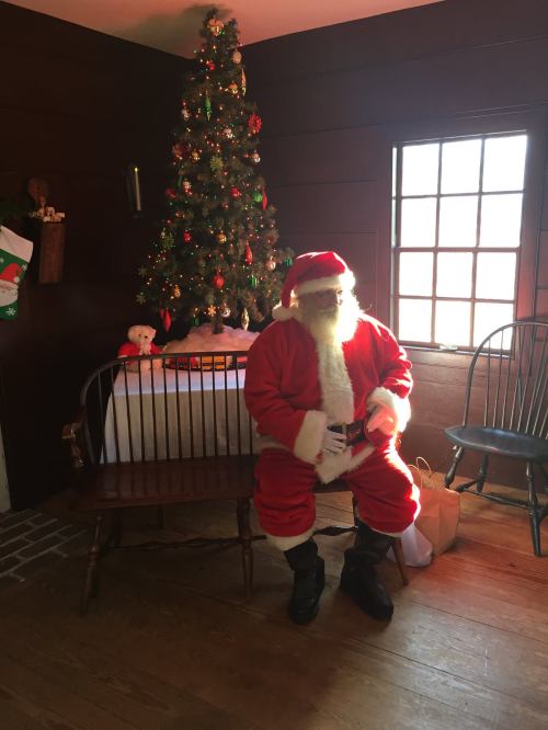 Santa Claus sits on a bench in a cozy room decorated for Christmas, with a tree and gifts in the background.