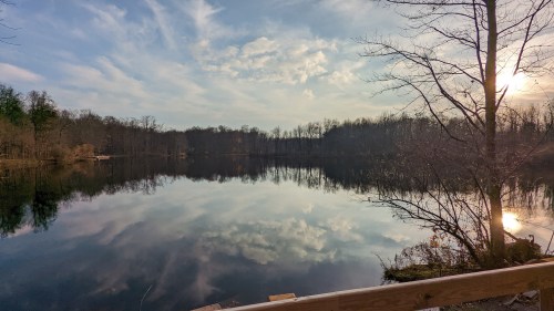 A serene lake reflecting trees and clouds, with a soft sunset glow illuminating the scene.