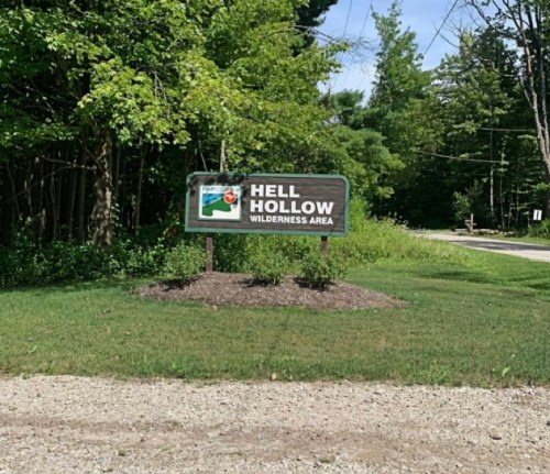 Sign for Hell Hollow Wilderness Area, surrounded by greenery and trees, near a gravel road.