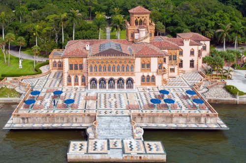 Aerial view of a grand mansion with a tiled terrace, surrounded by lush greenery and water.