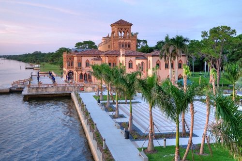 A scenic view of a waterfront building surrounded by palm trees and a walkway along the water at sunset.