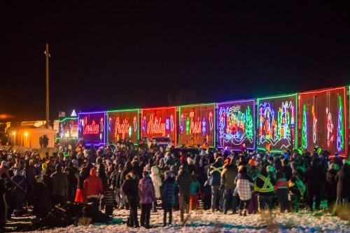 A large crowd gathers to watch a brightly lit train decorated with holiday lights at night.