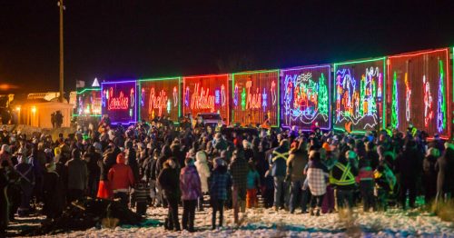 A large crowd gathers to watch a brightly lit holiday train display at night, surrounded by snow.