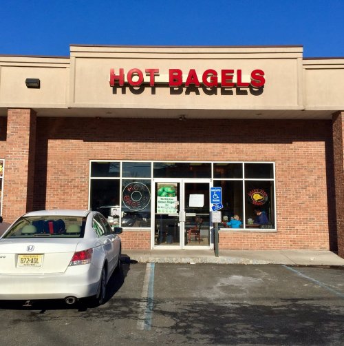 Exterior of a bagel shop named "Hot Bagels," featuring a brick facade and large windows, with a parked car in front.