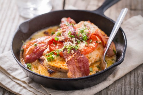 A cast iron skillet filled with grilled chicken, bacon, tomatoes, and herbs, served on a rustic wooden table.