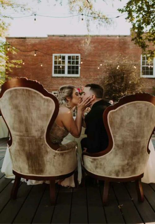 A couple shares a kiss while seated in elegant chairs at an outdoor wedding reception, surrounded by soft lighting.