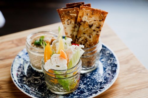 Three small jars filled with colorful vegetables and garnishes, accompanied by crispy crackers on a decorative plate.