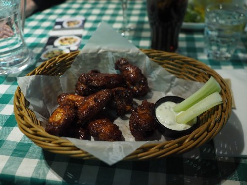A basket of crispy chicken wings with celery sticks and a small cup of dipping sauce on a checkered tablecloth.