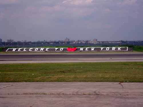 A large sign on a grassy area reads "WELCOME TO I ❤️ NEW YORK" with a city skyline in the background.