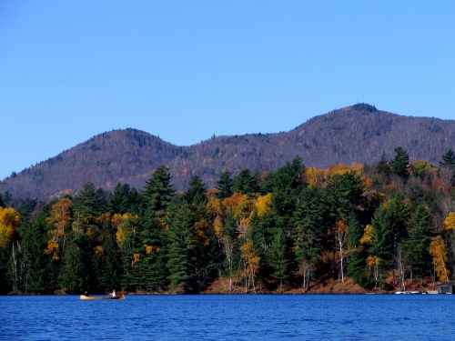 A serene lake surrounded by colorful autumn trees and mountains under a clear blue sky. A lone kayaker paddles peacefully.