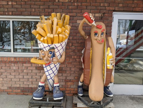 Two large, cartoonish statues: one of a smiling French fry cone and the other of a hot dog, both with playful expressions.