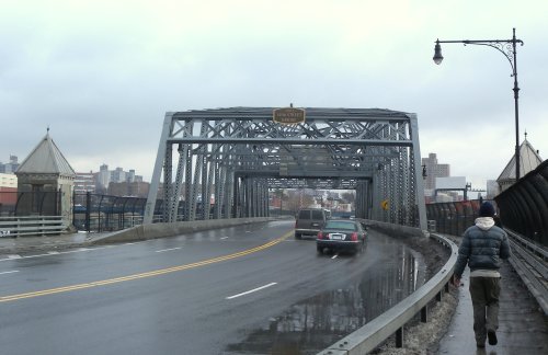 A gray metal bridge with a sign overhead, featuring a car on the road and a person walking along the side.