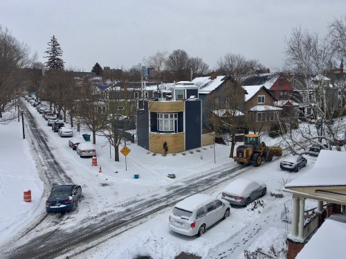 A snowy street scene with parked cars, a snowplow, and a modern house amidst winter trees and overcast skies.