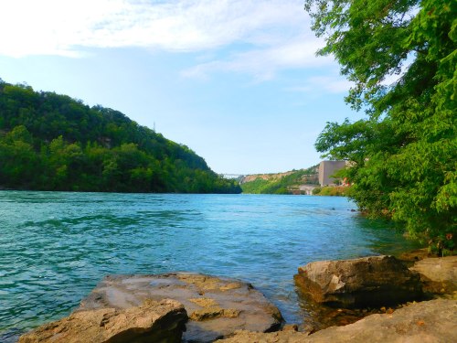 A serene river scene with clear blue water, rocky shore, and lush green hills under a bright sky.