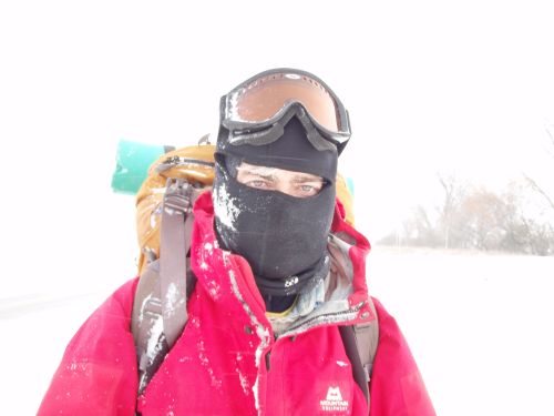 A person in a red jacket and goggles stands in a snowy landscape, partially obscured by a black face mask.