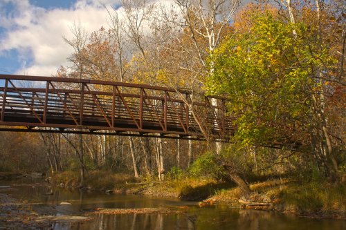 A metal bridge spans a calm stream, surrounded by autumn trees with colorful leaves under a partly cloudy sky.