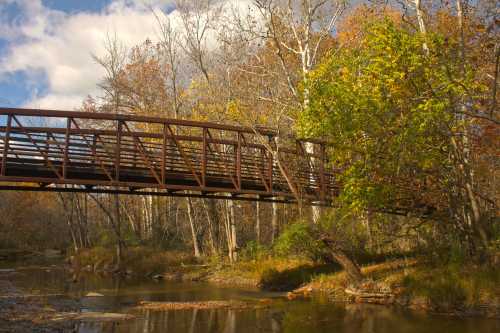 A metal bridge spans a calm stream, surrounded by autumn trees with colorful leaves under a partly cloudy sky.