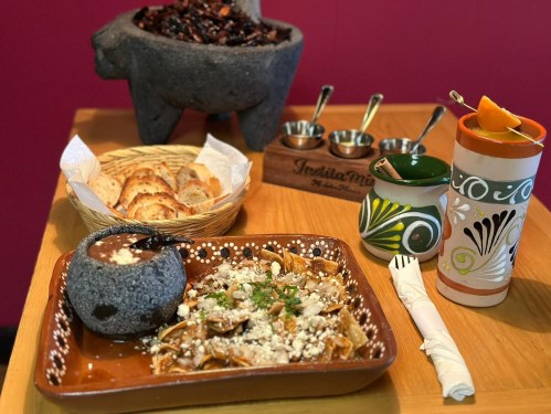 A colorful table setting featuring a traditional dish, bread, drinks, and a stone mortar, all against a vibrant background.