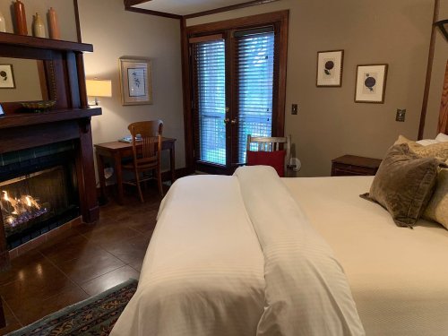 Cozy hotel room with a fireplace, a bed, a small dining table, and a door leading to a patio. Warm, inviting decor.