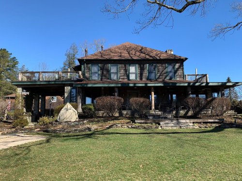A large, two-story house with a wraparound porch, surrounded by trees and a well-maintained lawn under a clear blue sky.