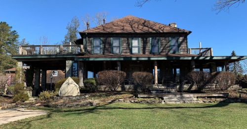 A large, two-story house with a wraparound porch, surrounded by trees and a well-maintained lawn under a clear blue sky.