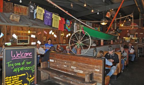 A rustic restaurant interior with wooden decor, a green canoe overhead, and patrons dining at wooden booths.