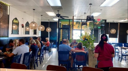 A busy restaurant interior with diners at tables, pendant lights, and a vibrant decor featuring plants and artwork.
