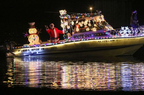 A decorated boat with Santa and a snowman, illuminated by festive lights, glides across a dark, reflective water surface.