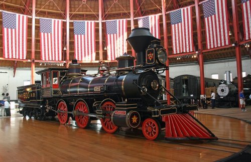 A vintage steam locomotive displayed in a roundhouse, surrounded by American flags and wooden flooring.