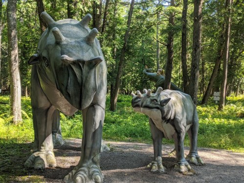 Two large dinosaur sculptures in a forested area, surrounded by trees and greenery.