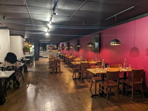 Interior of a restaurant featuring wooden tables, chairs, and pink walls, with pendant lights and a cozy atmosphere.