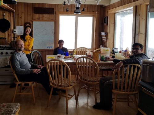 Four people sitting around a wooden table in a cozy cabin, with food and drinks spread out. Windows show a snowy view.