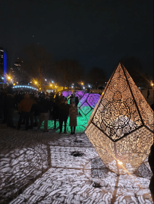 A nighttime scene with illuminated geometric sculptures casting intricate shadows, surrounded by a crowd in a snowy park.
