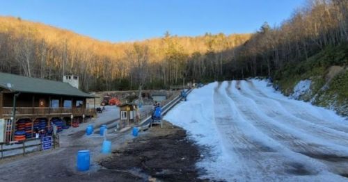 A snow tubing hill with multiple lanes, a lodge on the left, and trees in the background under a clear blue sky.