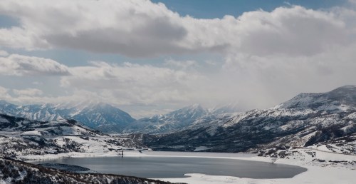 A serene winter landscape featuring a frozen lake surrounded by snow-covered mountains under a cloudy sky.