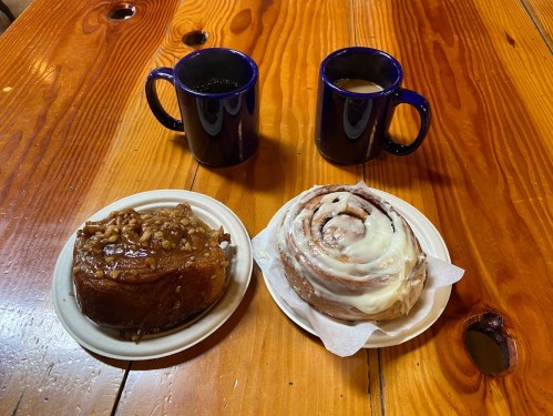Two mugs of coffee beside a plate with a sticky bun and a cinnamon roll on a wooden table.