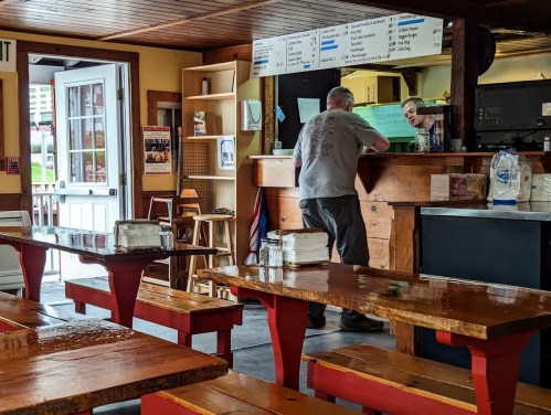 A man stands at a counter in a casual dining space with wooden tables and a menu board on the wall.