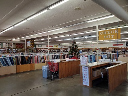 A spacious fabric store with rows of colorful fabrics, a checkout area, and a Christmas tree in the corner.