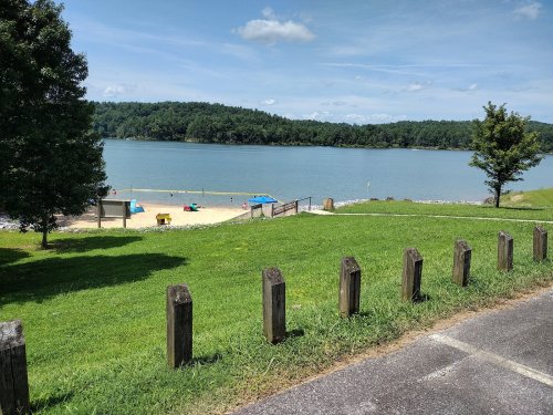A serene lakeside view with a sandy beach, green grass, and trees under a clear blue sky.