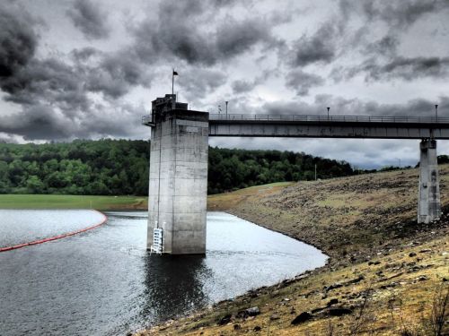 A concrete dam tower stands by a lake under a cloudy sky, with a rocky shoreline and a safety barrier in the water.