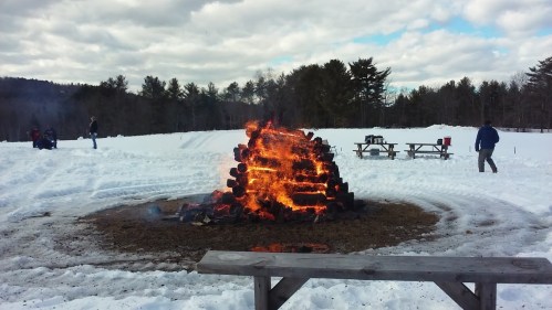 A large bonfire burns in a snowy landscape, with people in the background and picnic tables nearby.