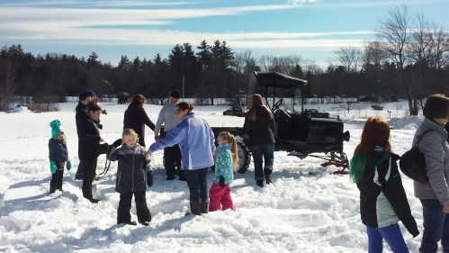 A group of people, including children, play in the snow near an old-fashioned vehicle on a sunny winter day.