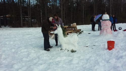 Two people sculpt a snow llama in a snowy field, while others build snow figures in the background.