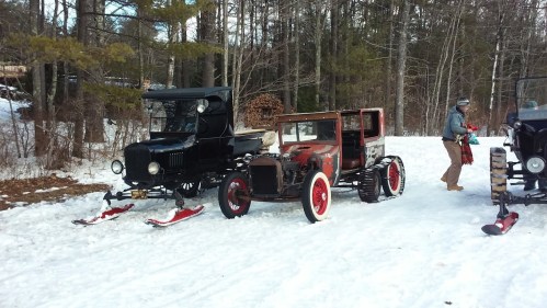 Two vintage cars with snow skis parked on a snowy landscape, surrounded by trees. A person is walking nearby.