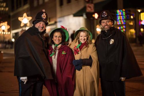 Four people in festive capes and hats pose together on a city street decorated with holiday lights.
