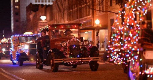 A festive parade featuring vintage cars adorned with colorful holiday lights on a city street at night.