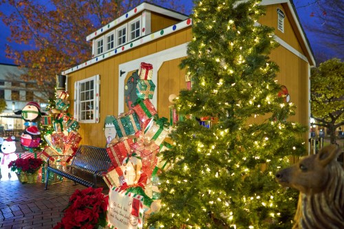 A festive scene featuring a decorated cabin, Christmas trees, and colorful gift displays, illuminated at night.
